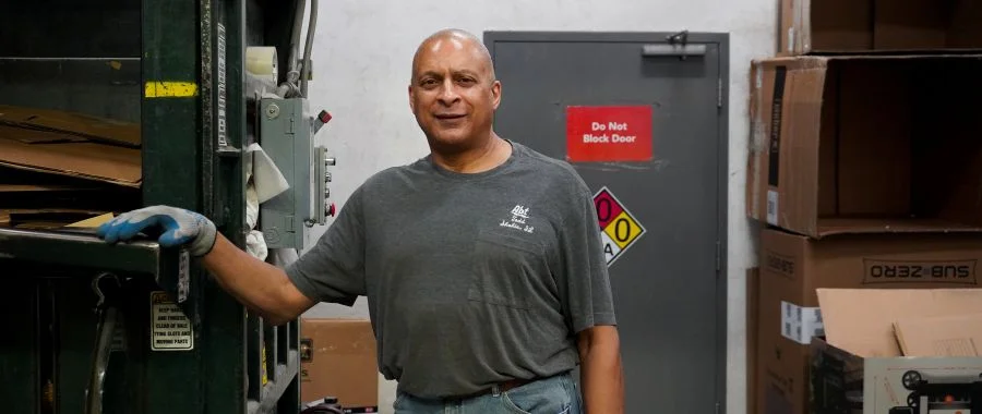 Todd Anderson in the warehouse, resting his hand on the cardboard crusher