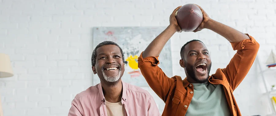 Two men watching sports and cheering. The man on the right is holding a football above his head.