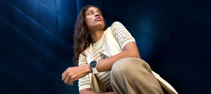 A woman wearing white against a dark blue background. She is wearing a white Garmin Venu 4 smartwatch.