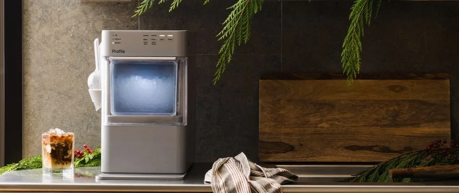 The GE Profile Opal Mini ice maker on a countertop decorated with holiday evergreen boughs beside a cup of iced coffee.