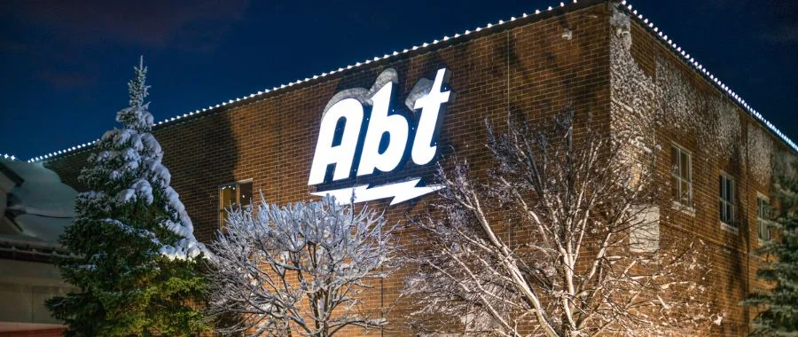 The outside of Abt Electronics at night with snow covered trees.