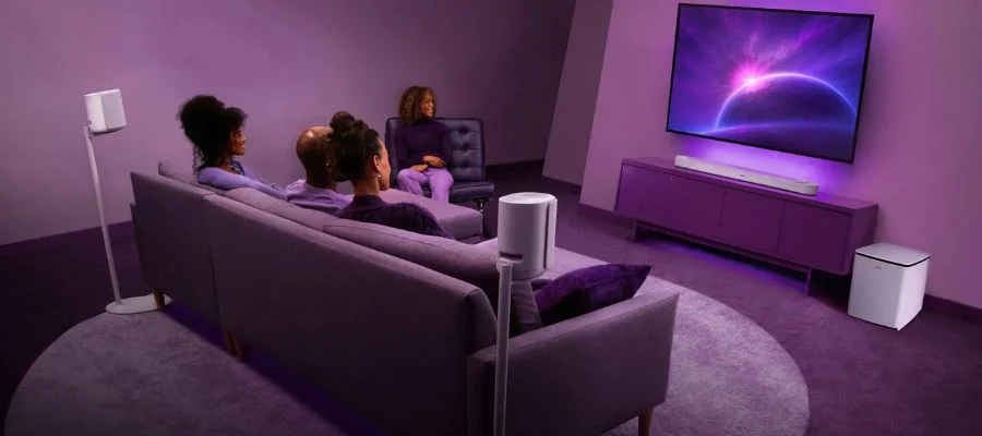 Four people sitting around a wall mounted TV with a Bose Lifestyle Ultra soundbar underneath, a Bose Lifestyle Ultra subwoofer and two Lifestyle Ultra speakers on stands.