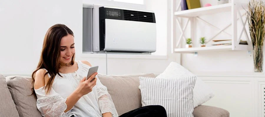 A girl sitting on a couch with an air conditioner in the window behind her.