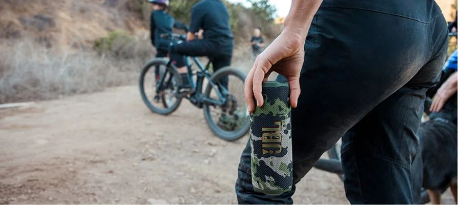 The JBL Flip 7 in a camo print. A person holds it by their side as priends prepare to bike along a dirt bike trail