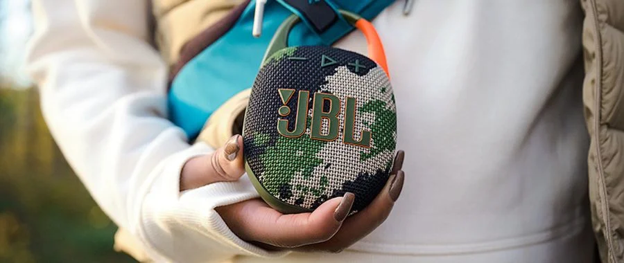 he JBL Clip 5 in camo is clipped onto a bag with a person holding it steady.