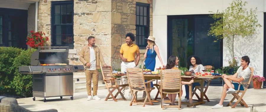 Group of people sitting at a table and around a grill on a sunny day.