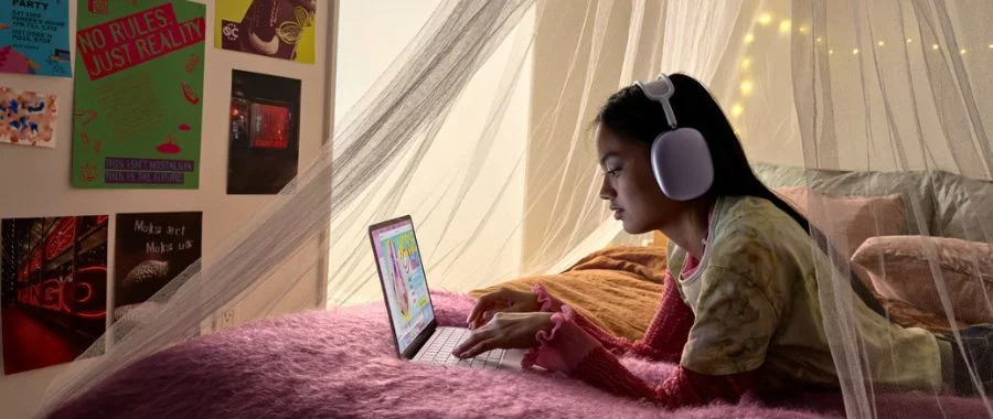 Young woman laying on a bed, wearing AirPods Max headphones and working on a MacBook Neo