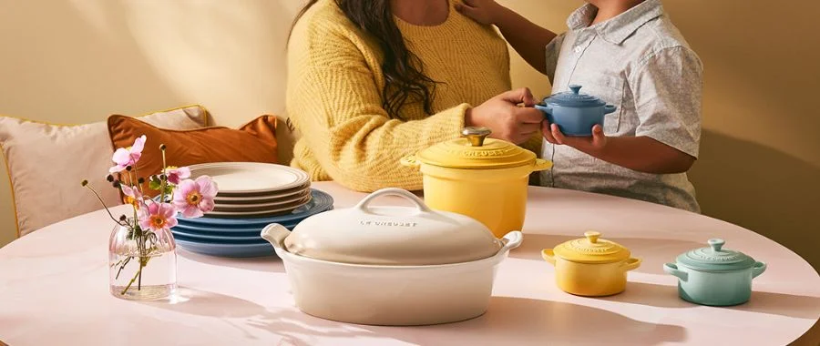Yellow, green and cream Le Creuset cookware pieces on a pink table. A little boy hands a small cocotte to his mom in the background.