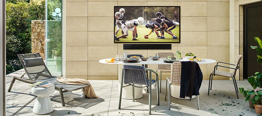 Samsung Terrace outdoor TV mounted on exterior wall over a soundbar. An oval dining table sits in front of the TV.