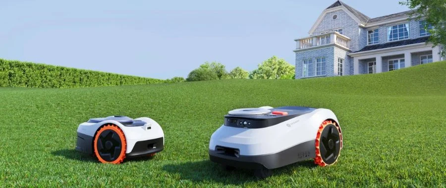 Two robot lawn mowers of different sizes on a large, neat lawn with a large brick house in the background.