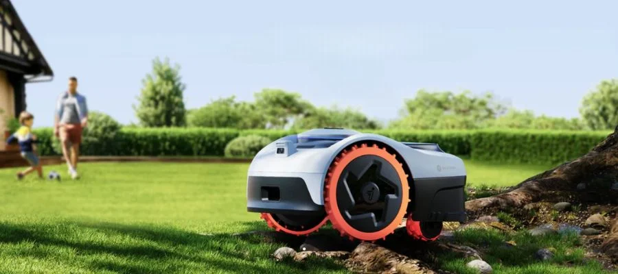 Robot lawn mower on the grass under a tree while a father and son play soccer in the background.