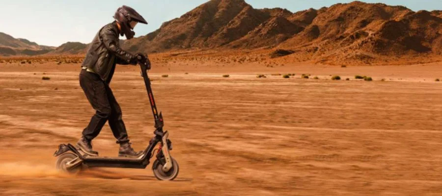 A person wearing a leather jacket, helmet and visor and riding a Segway scooter through an arid desert.
