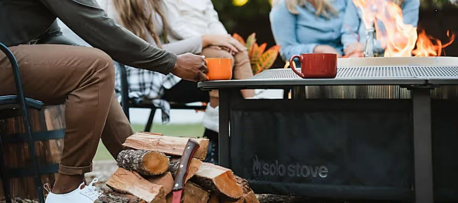 A close-up of a Solo Stove Surround protector holding up mugs of warm drinks beside firewood. Several people sit beside the fire