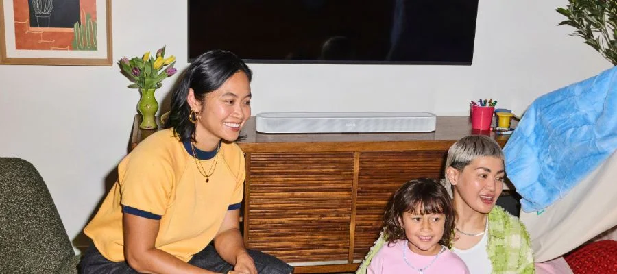 Two adults smiling with a young girl in front of a white Sonos Beam Ultra soundbar below a TV.