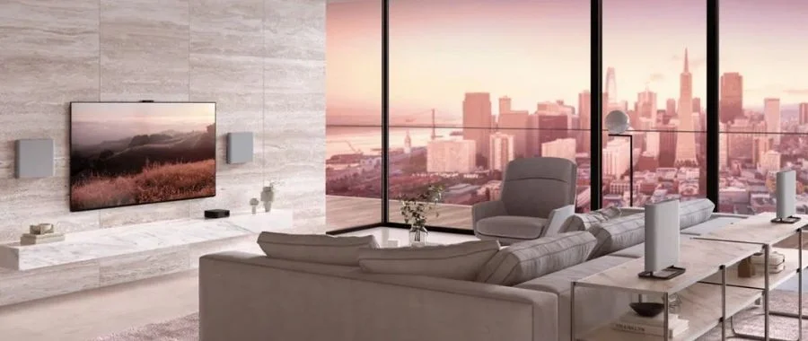 A Sony TV mounted on a wall with the two Bravia Theater Quad speakers mounted next to the TV and the other two speakers on a table behind a sofa opposite the TV.