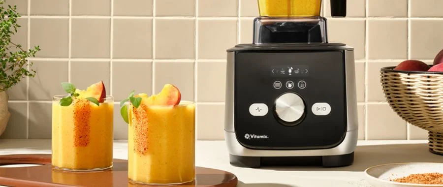 A Vitamix X Series blender on the countertop beside two glasses of a peach smoothie topped with peach slices.