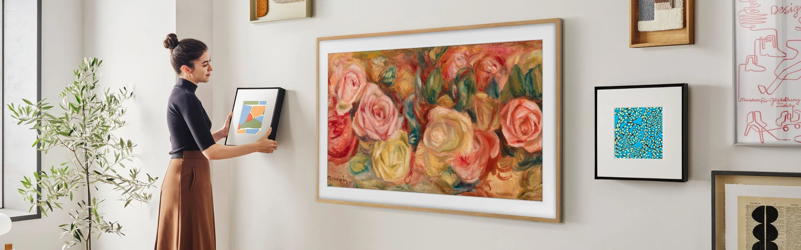 A woman hanging up frames around her art TV on a gallery wall. The TV is displaying a floral painting with a wooden or gold bezel.