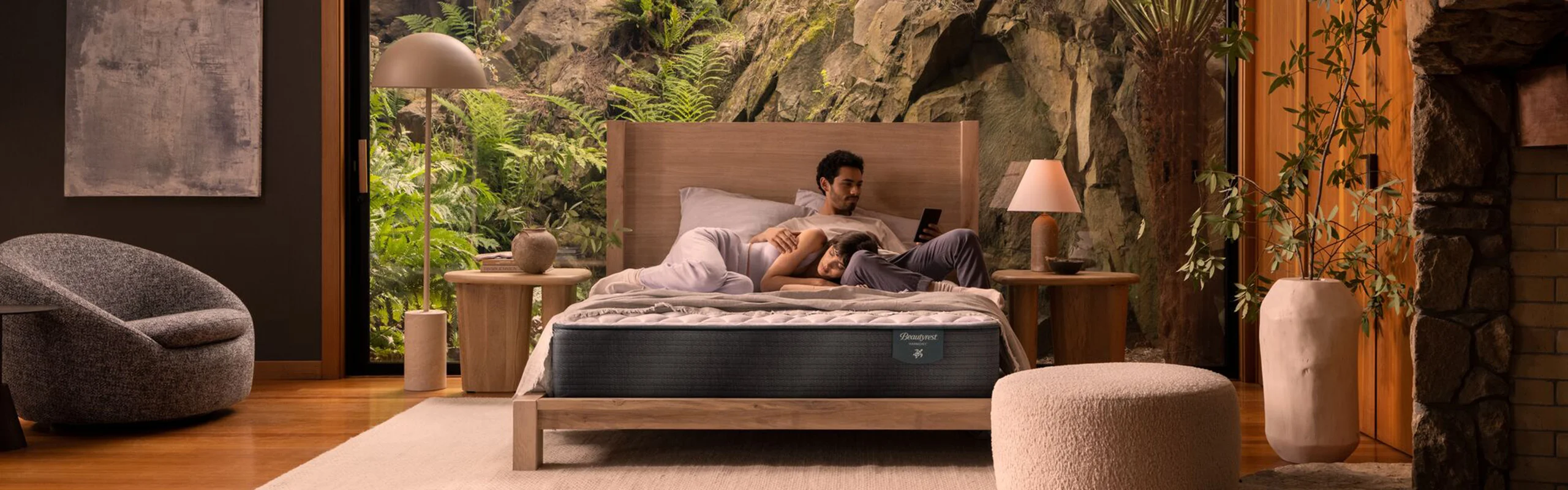 A couple relaxing on their mattress in a bedroom with a massive windo behind them, showing ferns and rock