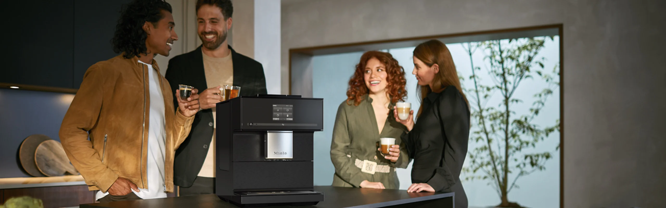 Four people with cups of coffee gathered around a kitchen counter holding a coffee maker