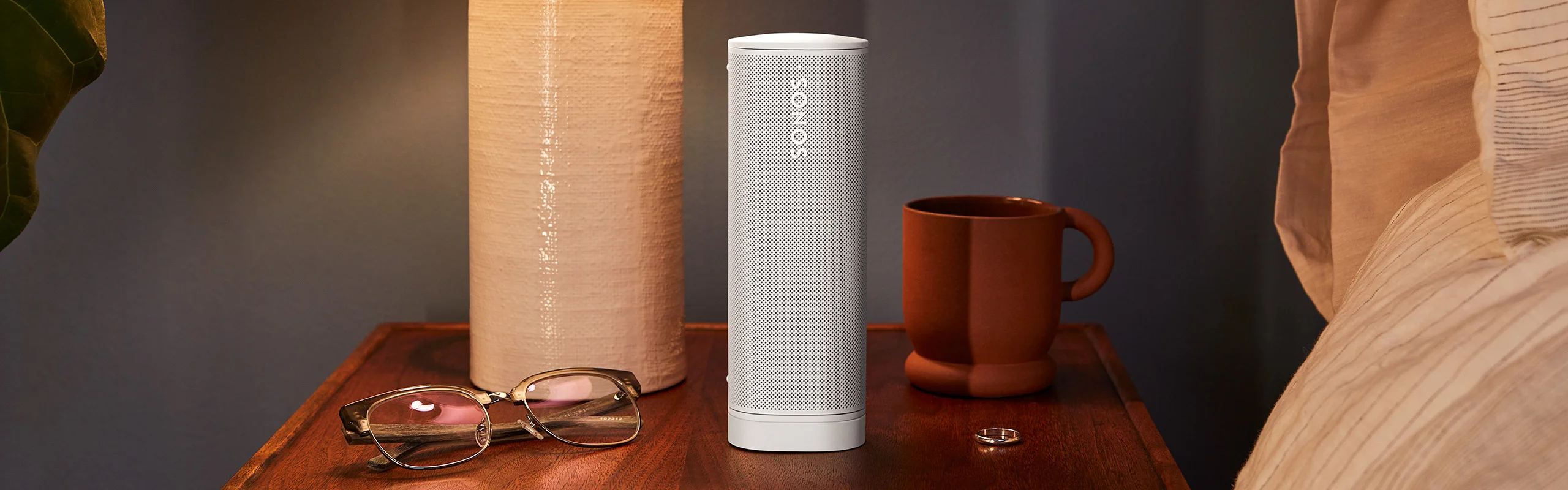A white speaker sitting on a nightstand next to a mug, a ring and a pair of reading glasses