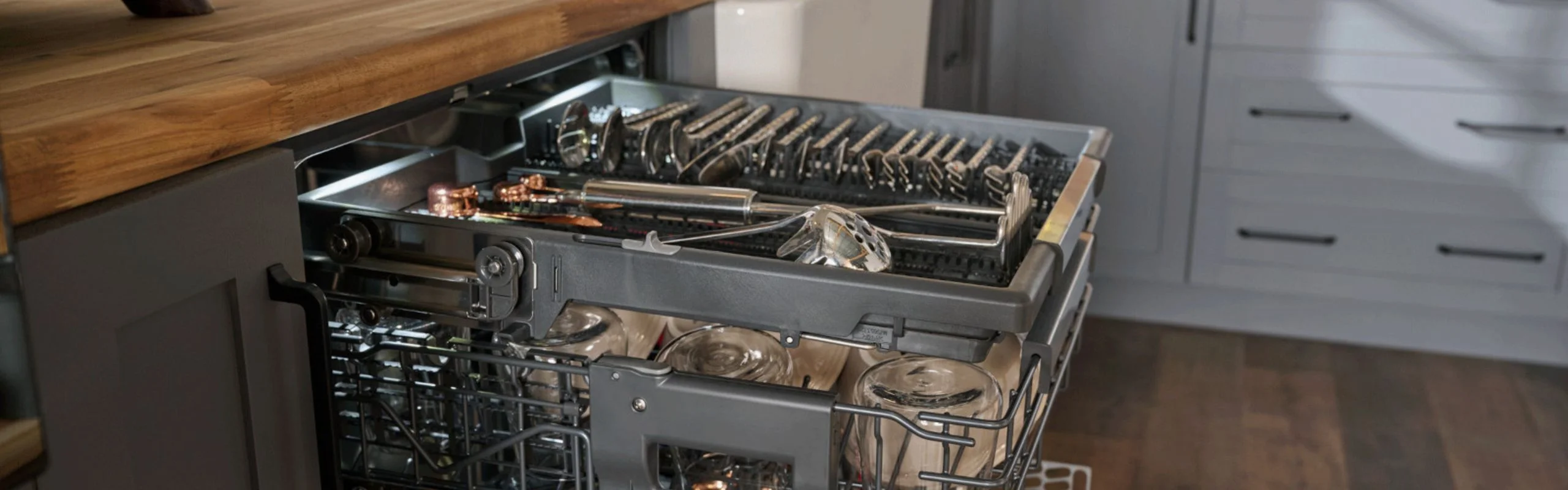 A dishwasher with a second and third rack rolled out, loaded with clean silverware and serveware in a gray kitchen.