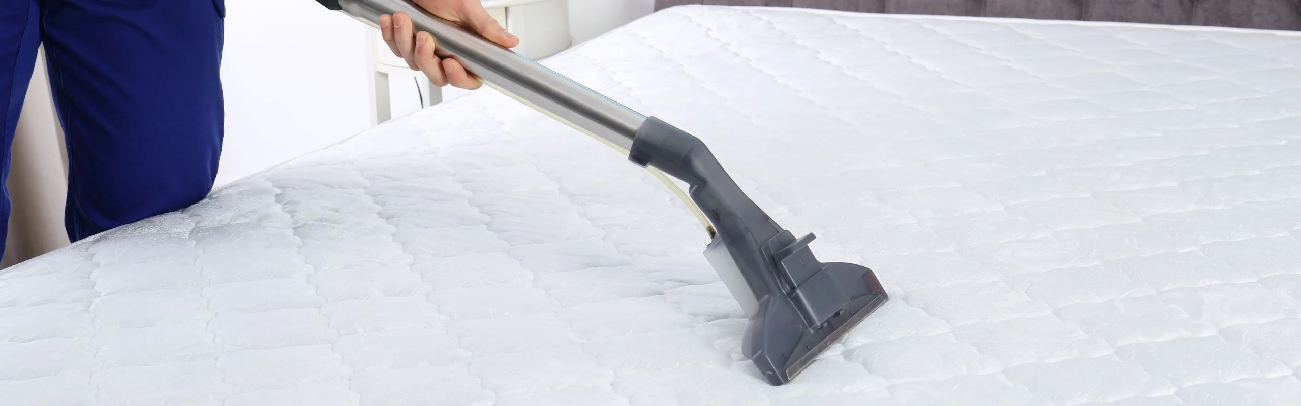A person using a vacuum and an upholstery attachment, using it to clean a white mattress.