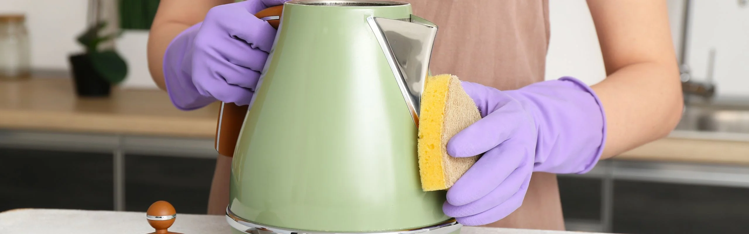 A person wearing gloves cleaning a green electric kettle with a sponge.